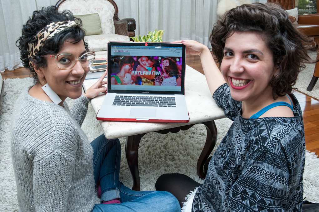 Two women are sitting on a white rug, looking over their shoulders at the camera while smiling and displaying the screen of an open laptop on a white marble coffee table in the center of the photo. The screen shows a photo of women carrying children and the word “Maternativa” in white letters. The woman to the left is wearing clear plastic rimmed glasses with large, round lenses, and white, rectangular earrings. She has short, dark brown wavy hair tied back with a leopard print headband, and is wearing a light gray sweater and jeans. The woman to the right has wavy, light brown, shoulder-length hair and is wearing a gray shirt with black print.
