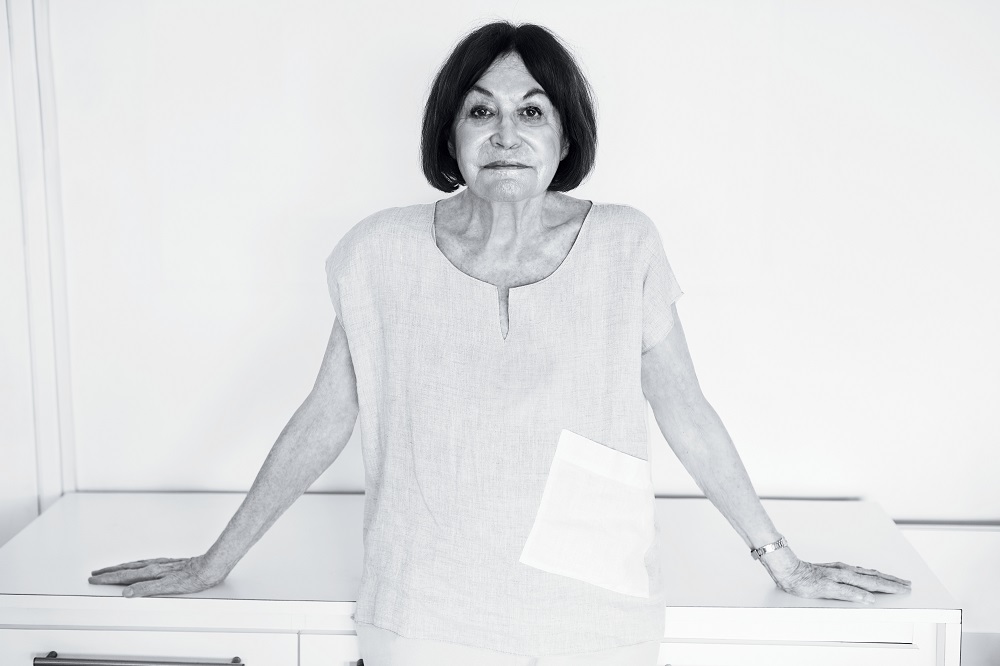 A black and white photo shows a woman with light skin, dark eyes, straight dark hair down to her shoulders, looking at the camera and resting her back and hands, her arms half open symmetrically, on a white cabinet. She is wearing a loose light colored shirt with short sleeves and a metallic bracelet on her left wrist. Behind her is a white wall.