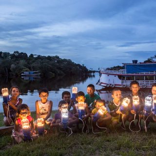 Several children are crouching on a lawn, facing the camera, while holding lamps built out of the lower half of plastic bottles, fitted onto blue electrical holders. In the background, on the right side, is a small boat anchored in a river, and a slightly cloudy sky at dusk.
