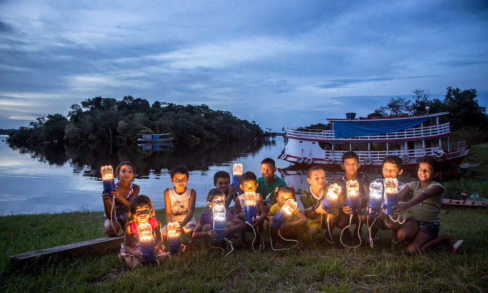 Several children are crouching on a lawn, facing the camera, while holding lamps built out of the lower half of plastic bottles, fitted onto blue electrical holders. In the background, on the right side, is a small boat anchored in a river, and a slightly cloudy sky at dusk.