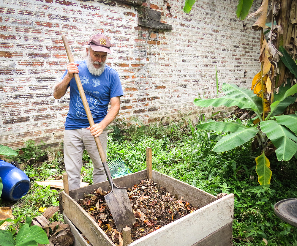 An older man with white skin and a full beard and moustache is holding a big shovel, with which he is working some dry leaves and dark earth in a square on the ground bordered by wooden slats. He is looking down at the earth. He is facing the camera and wears a brown cap, blue t-shirt and gray pants. Around him are bushes and low foliage. Behind him is a stained white brick wall.