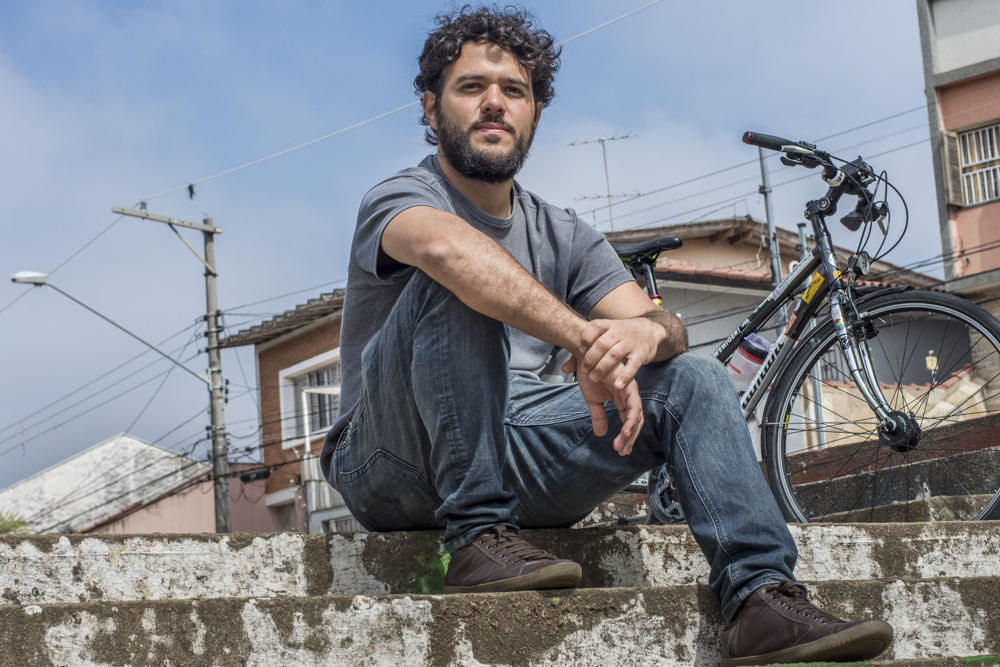 A man sits on gray concrete steps while resting his arms on his knees and looking at the camera. He has white skin, thin beard, short, wavy dark brown hair, and is wearing dark brown shoes, dark blue jeans and a gray t-shirt. Behind him, to the right, is the front wheel of a black and gray bicycle. In the background are houses, light poles and a bright blue sky with sparse white clouds.