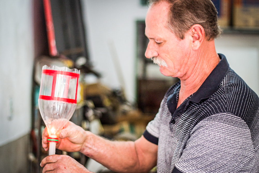 The same man from the cover photo, with white skin, gray hair and moustache, is facing the left side of the photo, holding the top half of a transparent plastic bottle that is cut in half vertically. He is wearing a short sleeved polo shirt with gradient stripes that go from white to black. The object he is holding has red tape shaped into squares around it. In the backgound are white walls with a variety of hanging objects out of focus.