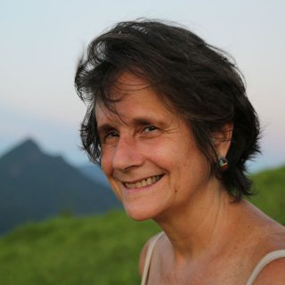 A woman facing the left side of the photo looks at the camera and smiles. She has white skin, straight brown hair, and wears a colorful circular earring and a white tank top. In the background, a valley of grassy ground and, in the background, the countenance of a hill and the blue-gray sky.