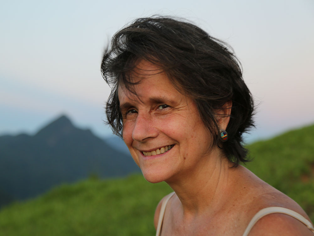 A woman facing the left side of the photo looks at the camera and smiles. She has white skin, straight brown hair, and wears a colorful circular earring and a white tank top. In the background, a valley of grassy ground and, in the background, the countenance of a hill and the blue-gray sky.