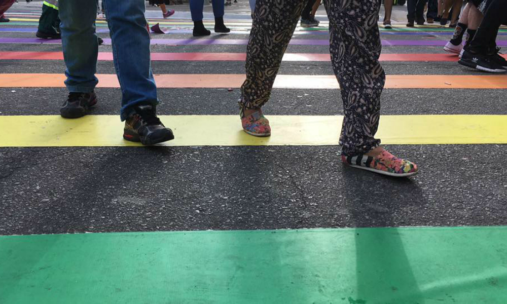 Os pés, calçados, de diversas pessoas, passando sobre uma faixa de pedestres colorida.
