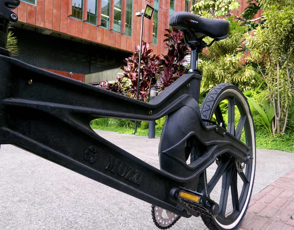 The photo shows the seat and back wheel of a black bicycle on a street, facing the camera diagonally from the left corner. In the back is a building covered with brick-colored tiles and, to the right, a garden with some short trees.
