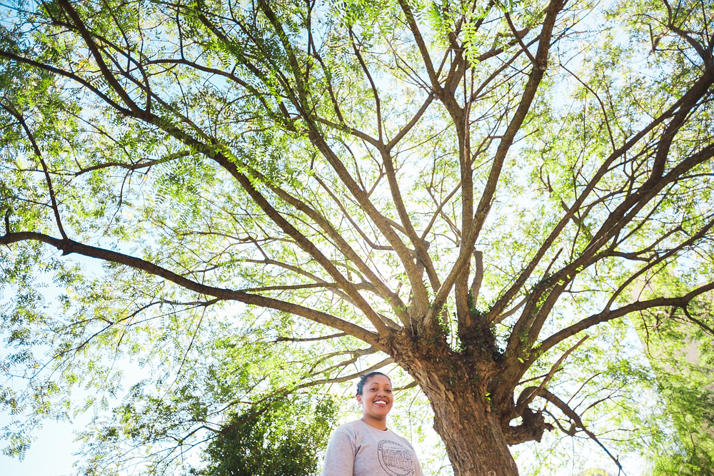 A smiling woman looks at the camera. She has dark skin and dark brown hair tied into a bun, and is wearing a light gray t-shirt with a black circular design in the middle. Behind her is a big tree with light green leaves and branches spreading out, and the blue sky behind it.