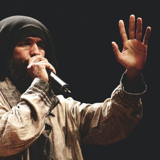 A man with brown skin, brown curly beard and a black cloth covering his hair talks into a microphone. He is wearing a loose light brown shirt made of light cotton, and is facing diagonally towards the right side of the photo, his left hand held up and open, showing his palm. His right hand holds the microphone up to his mouth. The background is black.