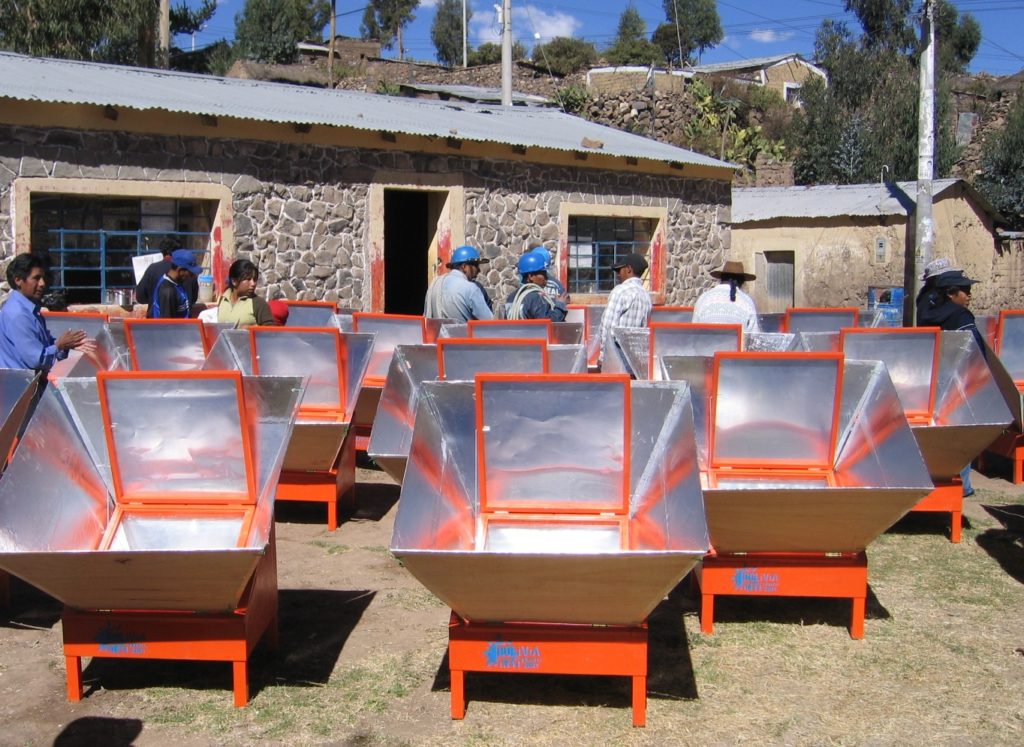A foto mostra mais de 10 fogões solares em um espaço aberto, no meio de um conjunto de casas feitas de pedra (que vemos ao fundo). No meio destes fogões (que se parecem a caixas de madeira e alumínio), vemos umas 10 pessoas ao fundo, de costas, usando capacete de proteção.