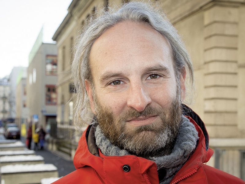A man with gray hair and beard, 46 years old. He wears a gray wool sweater under a red winter coat. In the background are buildings and a street.
