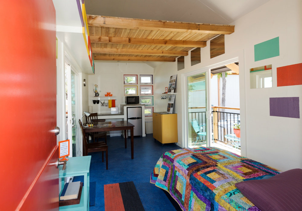 ALa foto muestra el interior de una pequeña casa, donde vemos una cama, al fondo una pequeña cocina, con una mesa, dos sillas, microondas y refrigerador. Al lado un pequeño balcón. Las paredes y la decoración tienen muchos colores.