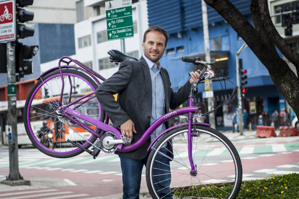 A brown-haired, lightly-bearded, white man wearing jeans, a light blue shirt and a dark gray blazer holds a purple bike. In the background is an intersection with bike paths, pedestrian lanes and a municipal bus.
