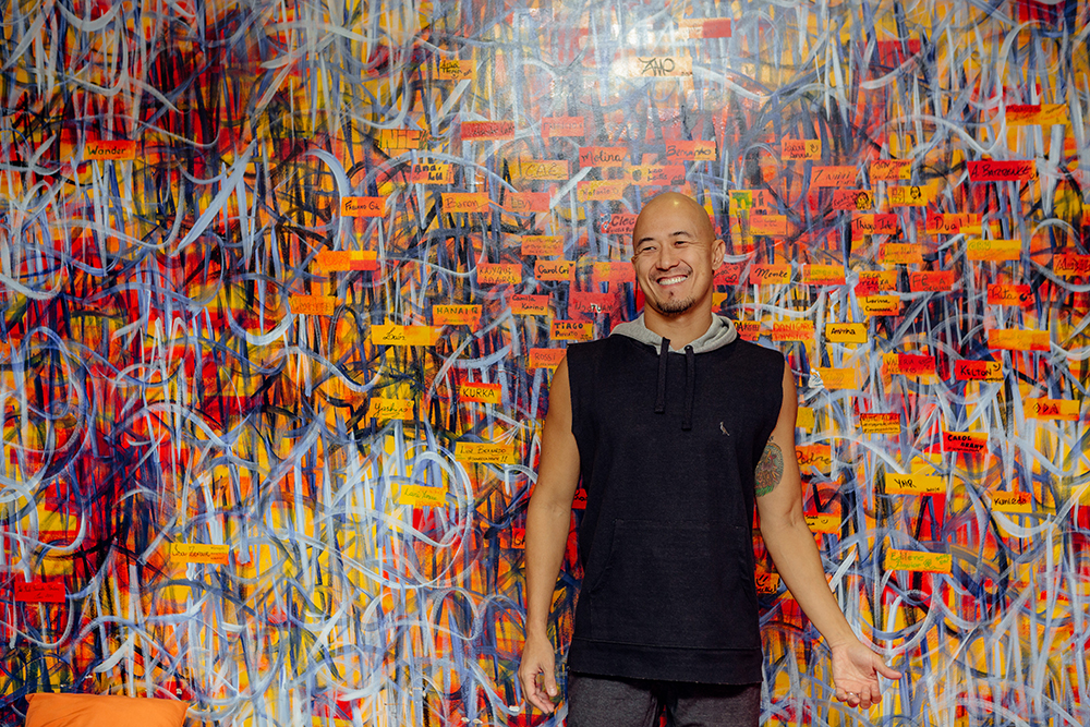 A bald, Japanese man with a small beard. He wears a blue shirt and dark gray pants. He is looking to his right side and smiling. There is a wall painted colorfully with graphite in the background.