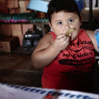A chubby boy with short black hair, brown eyes and dark skin is eating some potato chips, his hand to his mouth. He looks at the camera, holding up the blue packaging of the snack.