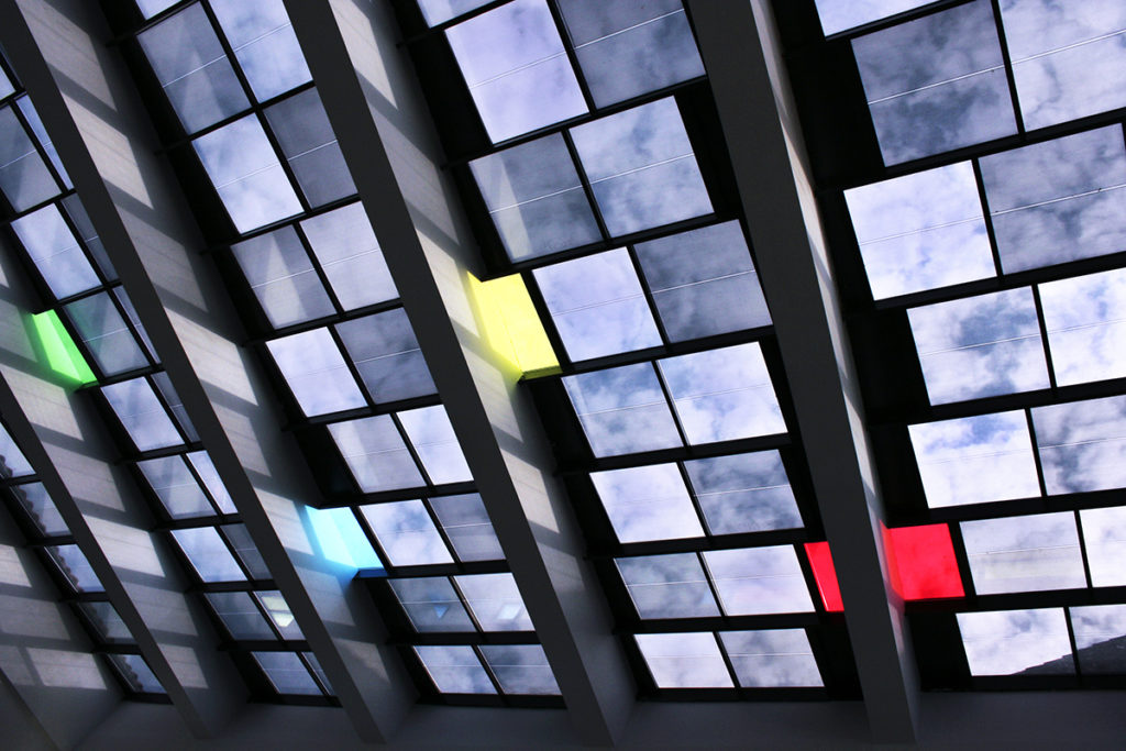 A close-up of glass cut into squares, with clouds and blue sky reflected in most of them. Three stand out, reflecting something else: the colors red, yellow and green. Each group of 15 glass squares is separated by a steel beam.