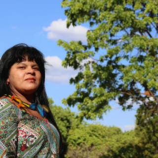 A middle-aged indigenous woman with long black hair and bangs, wearing a printed blouse. There are trees in the background.