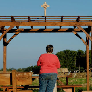 The picture shows a wooden open-air structure. There is a cross with a heart symbol on the roof. In the foreground there are some wooden benches. A woman with short brown hair wearing jeans and a long-sleeved pink shirt, her back to the camera, faces the wooden structure. She is praying. In the background, an open green area and some trees, all under a blue and cloudless sky