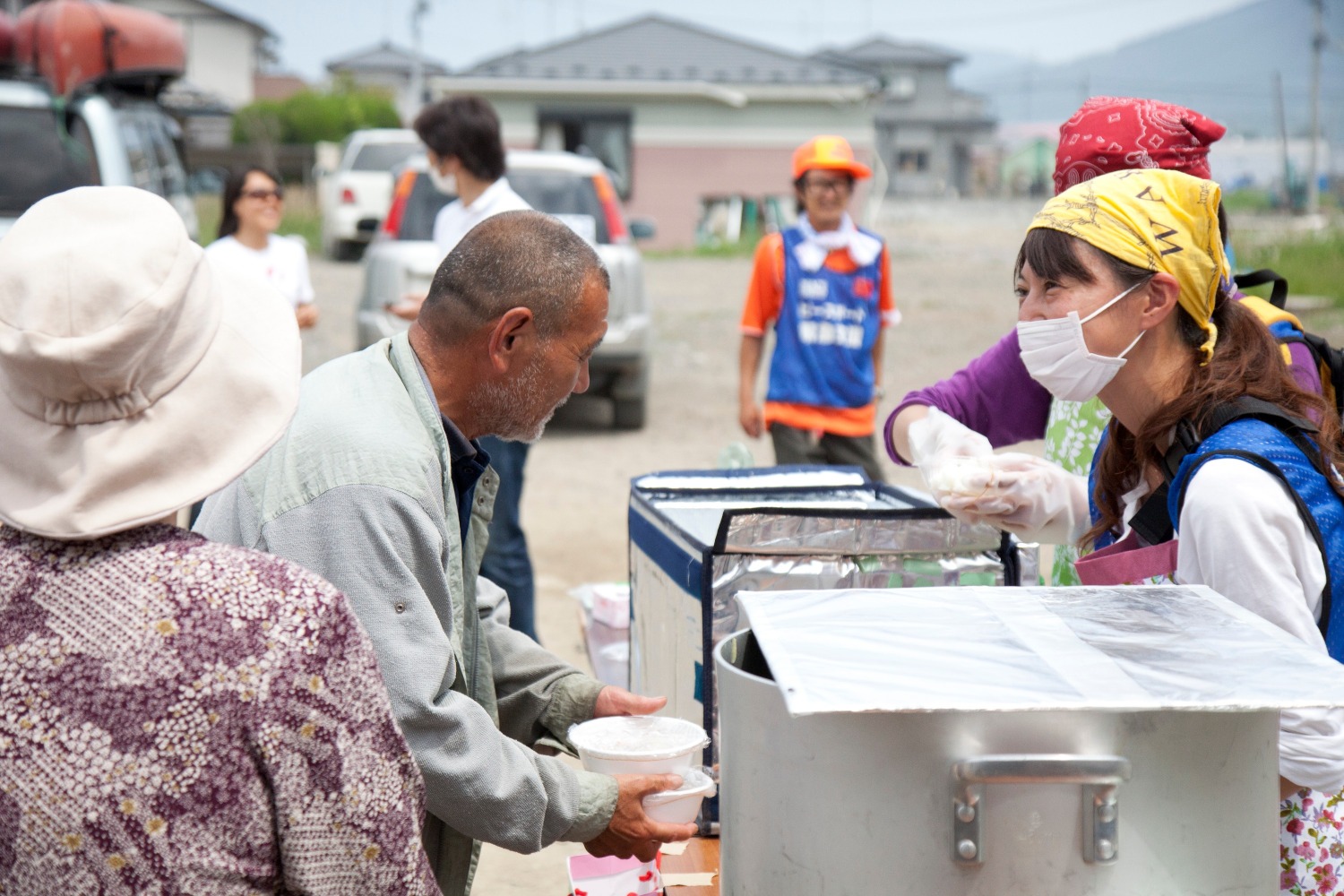 A young Japanese woman wears a mask covering her mouth and nose. She smiles and stands behind a table, which holds a large pot used to cook food. A man with a gray crew-cut and a gray beard looks at her, holding a couple bowls in a sign of respect and thanks.