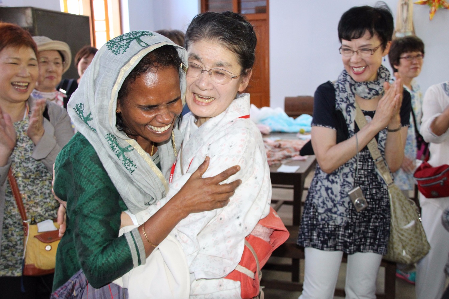 A Japanese woman in her sixties, wearing a white tunic, hugging a black-haired woman who is wearing a green Indian dress, and, on her head, a white scarf embroidered with a tree pattern. Both women smile spontaneously. Around them, other women smile and clap their hands.