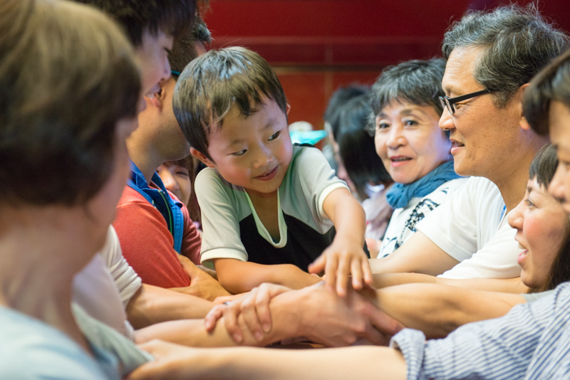 Two rows of people face each other. Each one holds the arm of the person in front of them. In the middle, leaning on these intertwined arms, a Japanese child, age around 4 or 5, looks at one of the adults.
