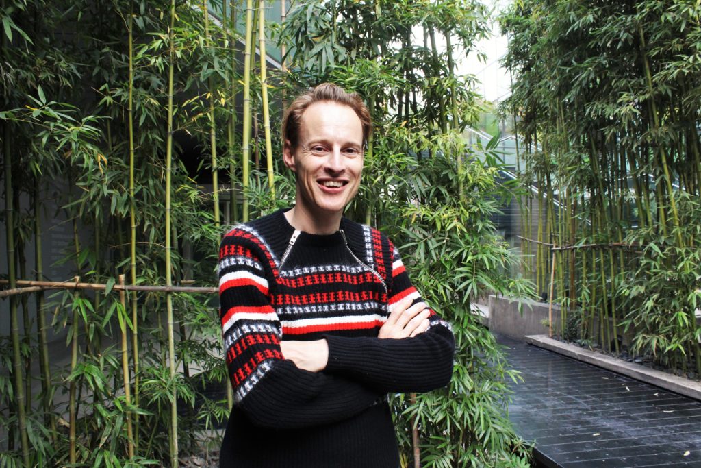 A thin, white man with straight, blond hair is smiling at the camera. He is standing with his arms folded. He wears a black wool sweater with red and white stripes across the chest. In the background is part of a walkway lined with trees.