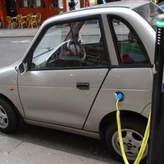 On a street, a small, silvery-colored electric car plugged with a cord into a pole. On the pole is written, “City of Westminster, Elektrobay Electric Vehicle Recharging Site.”