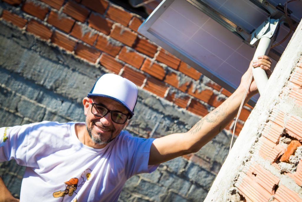 A brunette man, wearing a white T-shirt, a blue and white cap, and glasses with black rims, looks directly at the camera and smiles. His left hand rests on the arm of a photovoltaic panel, which is attached to a building’s exterior. His other hand is at his waist. Behind him is an exposed brick and concrete wall.