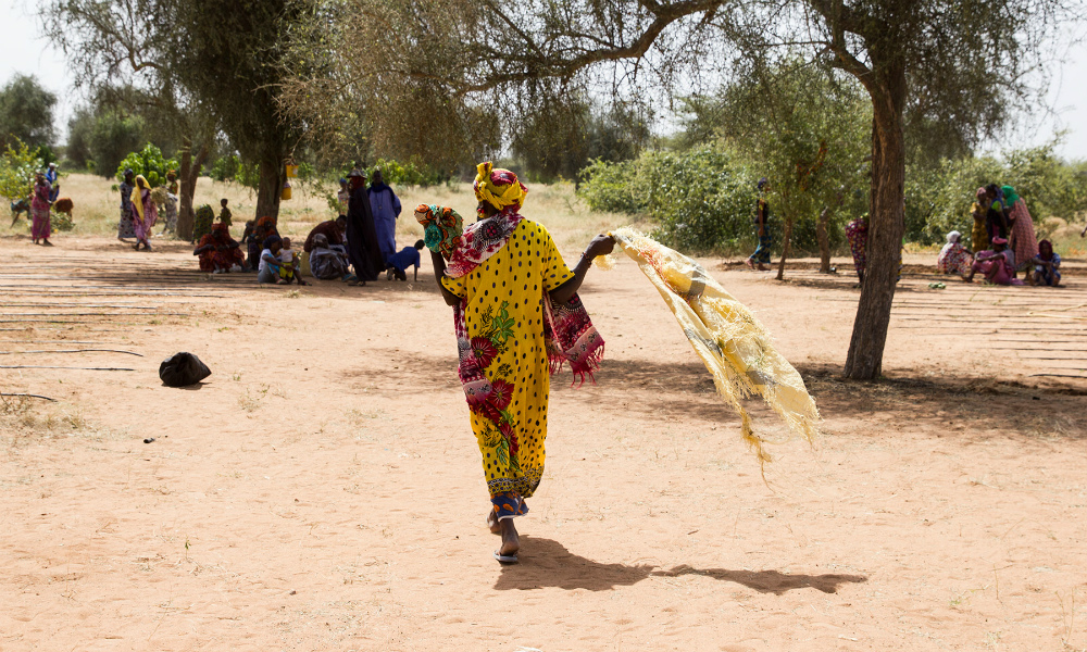 A black woman is wearing a long dress and a colorful turban (yellow with a red and green print), in the middle of the image, her back to the camera. She holds a yellow scarf and walks toward a group of about 15 black people gathered in the shadow of a tree. The land is desert, with sand and few trees.