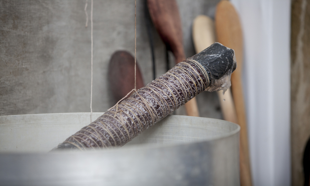 Primer plano de un trozo de palo de caña, donde está envuelto un hilo de color marrón. Atrás, aparecen cuatro cucharas de madera fuera de foco.