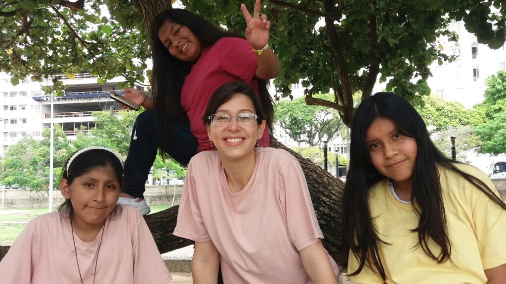 A woman with her hair tied back, wearing glasses and a pink t-shirt, is surrounded by three teenage girls, one of whom is sitting in a tree behind the woman, making a peace gesture with her fingers.