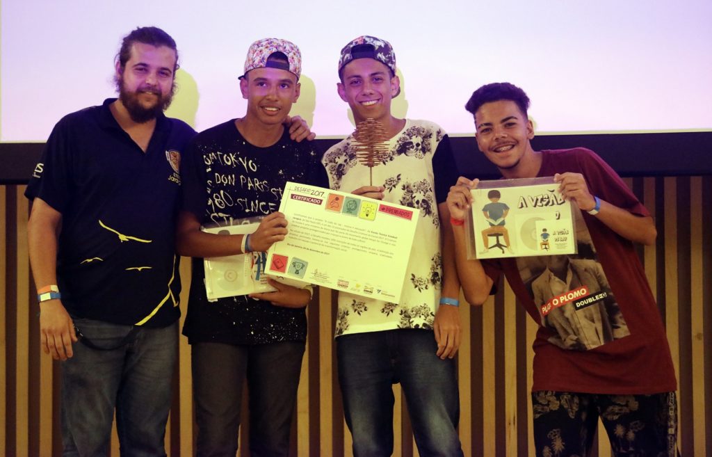 The photo shows a white man with a beard, his hair tied back, wearing a black t-shirt and jeans, next to three teenage boys. The first boy is wearing a patterned baseball cap, a black t-shirt with words printed on it in white, and jeans. He is holding the Design for Change certificate. Next to him is another teenage boy. This one is wearing a patterned baseball cap, a white t-shirt with a black pattern, and jeans. The last boy, on the right, wearing a brown t-shirt and black pants, is smiling and holding the "Viewed through Rap" curriculum.