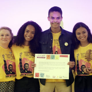 Four people stand side by side, each wearing a yellow t-shirt showing an image of a black man holding up his right fist. On the left end is an older white woman with light brown hair. Next to her is a young black woman with long curly hair. Beside her is a young black man, much taller than everyone else in the photo, holding a certificate. And on the right end is a young black woman with straight, dark brown hair, wearing hoop earrings.