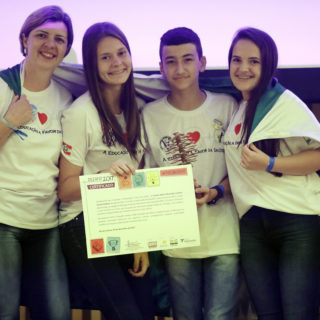 The photo shows four people, all wearing jeans and white t-shirts with the same design. On the left is a white woman with short light brown hair. To her right, a teenage girl with long brown hair holding a certificate. Next to her, a teenage boy with short dark hair. And on the right is a teenage girl with long dark hair. All four of them are wrapped in a green and white flag.