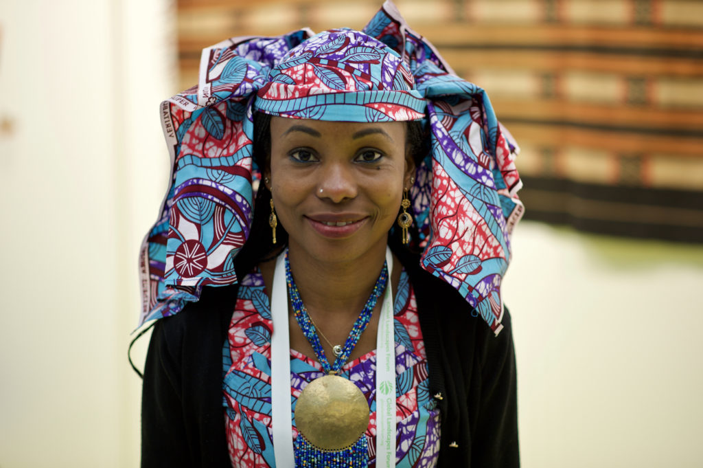 A black woman is wearing a blue necklace with a large golden medallion and, on her head, a blue, purple, red and white print scarf. She is also wearing a shirt or dress that matches her colorful scarf, and over it, a black sweater. She is smiling at the camera. She is in the center of the photo, with the background out of focus.