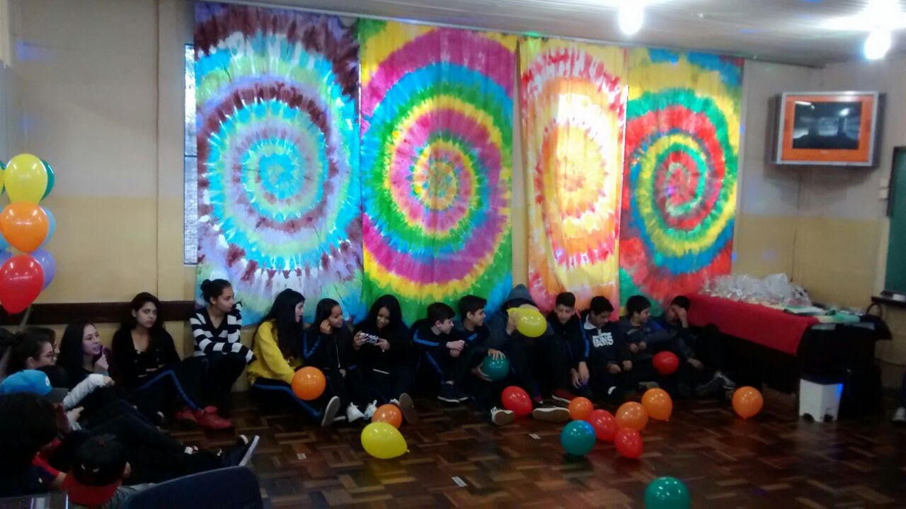 A group of teenagers sit on the floor of a classroom. The walls of the room are decorated with colorful banners and balloons are spread over the floor.