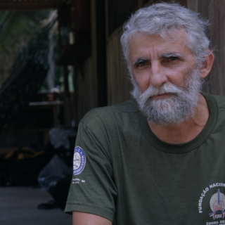 A man with gray hair and a beard, wearing a dark green t-shirt, looks at the camera. In the background is a wooden house and some of its interior.