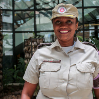 A black woman, her hair tied back, wearing a beige police uniform (a beige cap and matching jacket). She smiles at the camera.