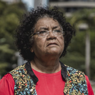 An elderly black woman with short curly hair, wearing glasses, a red T-shirt and a colorful, flowered vest, shown from the bust up. She is looking up slightly. The background, trees and concrete buildings, is out of focus.