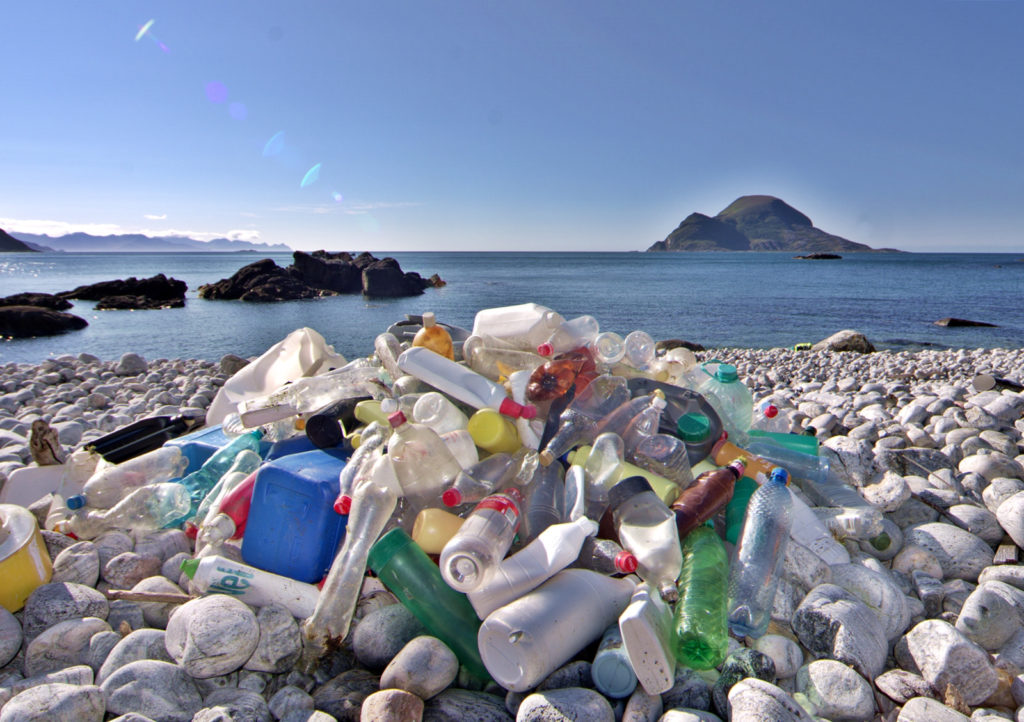 Plastic bottles and other plastic waste are piled up on white stones on a rocky beach, with the sea and a small island in the background.