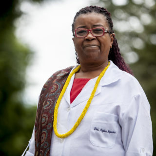 A black woman with long, braided hair, tinted red, wearing red-rimmed glasses, is looking at the camera with a faint smile. She wears a white lab coat and under it, a red T-shirt. She wears a dark brown, patterned scarf over her right shoulder, and a yellow necklace.