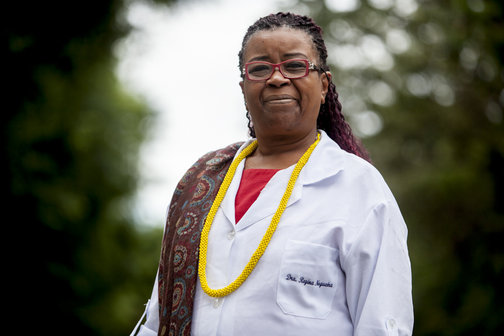 A black woman with long, braided hair, tinted red, wearing red-rimmed glasses, is looking at the camera with a faint smile. She wears a white lab coat and under it, a red T-shirt. She wears a dark brown, patterned scarf over her right shoulder, and a yellow necklace.