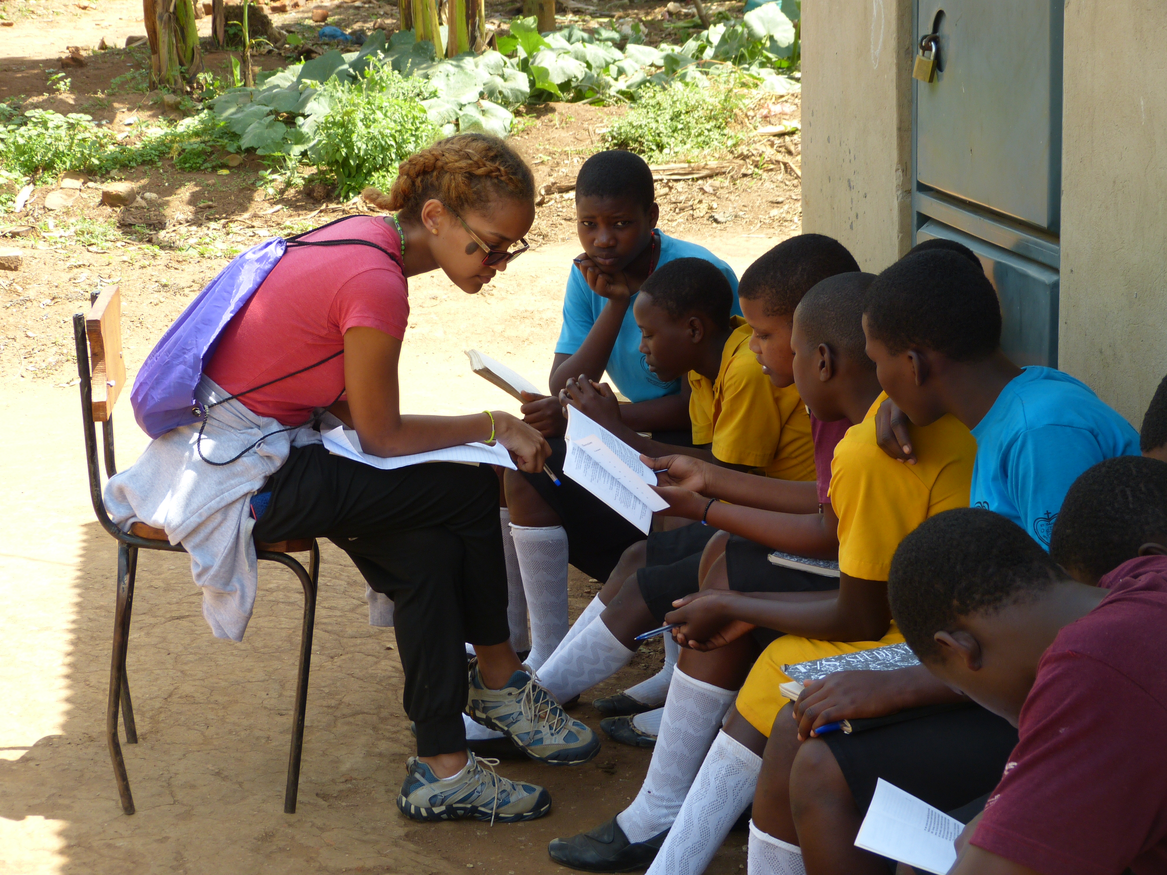 A young black woman with light-colored hair is wearing a pink T-shirt, black pants, sneakers, glasses, and a small cloth backpack. She is sitting on a chair, teaching a group of eight black boys, who are all sitting side by side looking at books. Some are also holding composition notebooks. The lesson is taking place outdoors, on a bench, and there are some vegetables growing in the background.