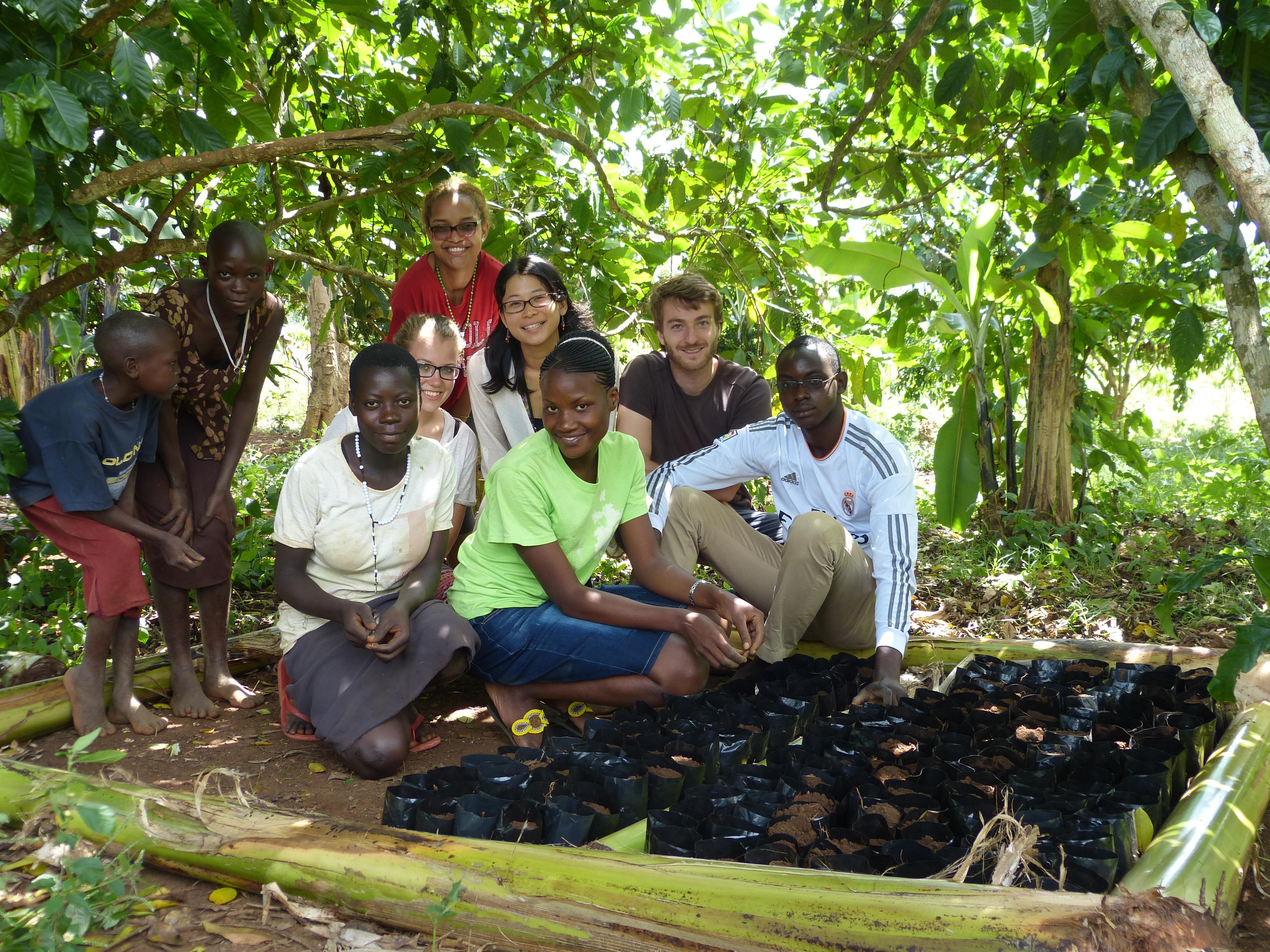 A group of nine people, almost all looking at the camera. They are gathered beneath some trees, sun shining through the leaves. Some are smiling, some serious. On the left, two black children stand a little bit apart from the group. In the center of the image are seven young adults, crouching or seated. In front are two black women wearing short skirts and a black man wearing a soccer jersey. Just behind them, a blonde, white woman, an Asian woman, and a blond, bearded man. In back of them is a young light-skinned, light-haired black woman wearing sunglasses and a red t-shirt. Next to the group, about a hundred seedlings.