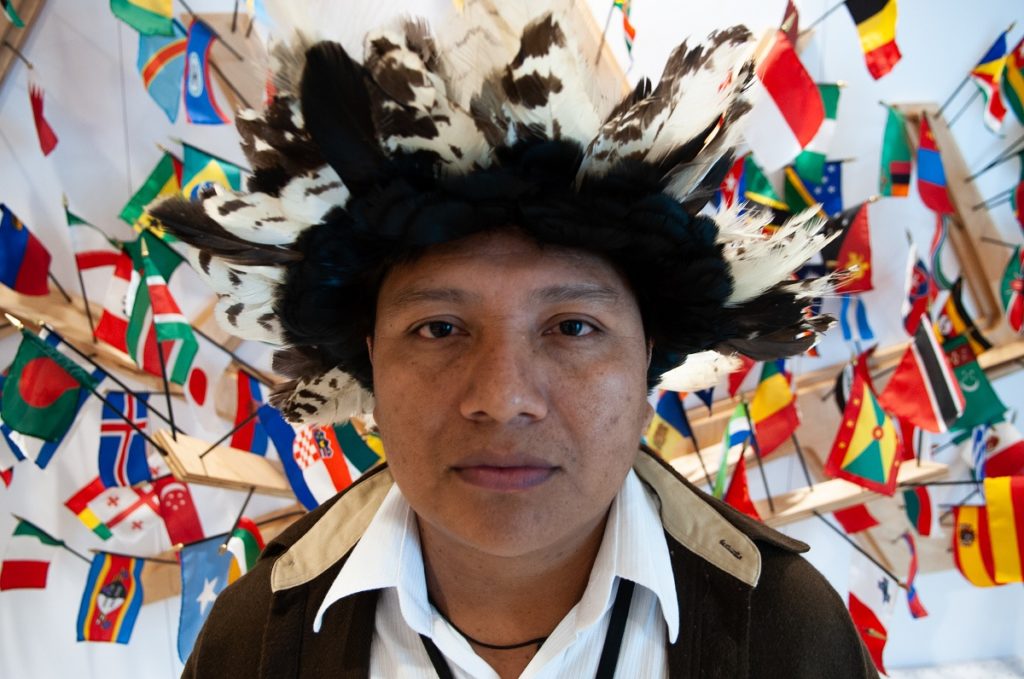 Hombre indígena, 32 años, piel morena y ojos oscuros, con una toca (cocar) de plumas negras y blancas en la cabeza, mira a la cámara, sin sonreír. Detrás, hay pequeñas banderas de varios países fijadas en la pared. La imagen le muestra de hombros para arriba.