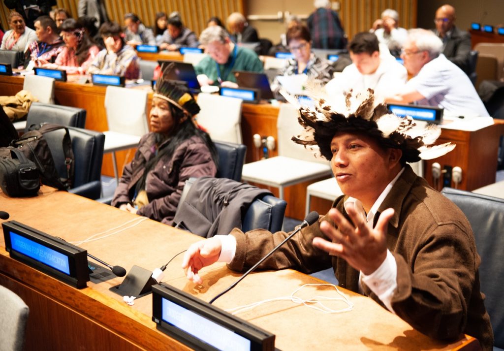 An indigenous man, 32 years old, with dark skin and black eyes, wearing a headdress made of white and black feathers, is sitting at a table in the United Nations in the lower right corner of the image. He is wearing a brown jacket with a white shirt underneath. He is speaking and gesturing with his right hand. Sitting next to him is an indigenous woman wearing a headdress and a gray jacket, looking straight ahead. Behind them are more people out of focus.