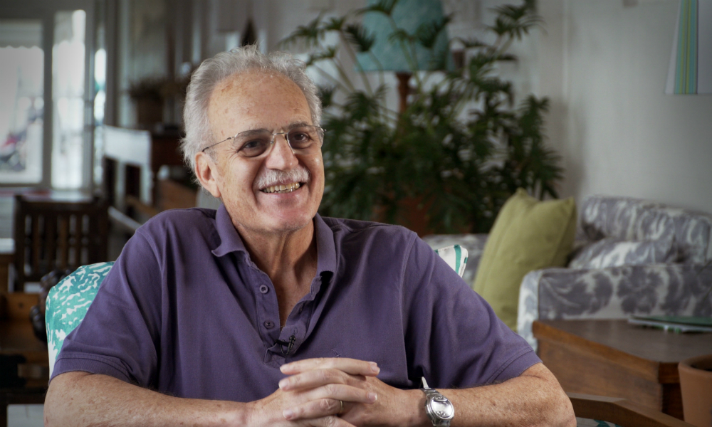 A 66-year-old man with a mustache and white hair, a bit bald. He wears glasses and a short-sleeved purple polo shirt. His forearm is resting on a table and his hands are clasped together. He smiles, looking to his left. In the background is a living room with a sofa, a large wooden table and a large, leafy green plant.