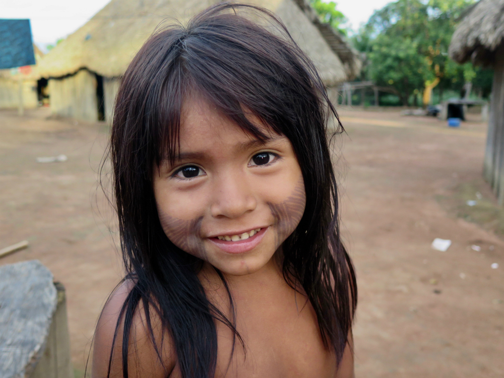 An indigenous girl, who looks about 7 years old, with big black eyes and straight black hair down to her chest. She is looking at the camera and smiling slightly. The lower halves of both of her cheeks are painted with black stripes. Behind her, shown only partially, is a wooden house with a thatched roof.