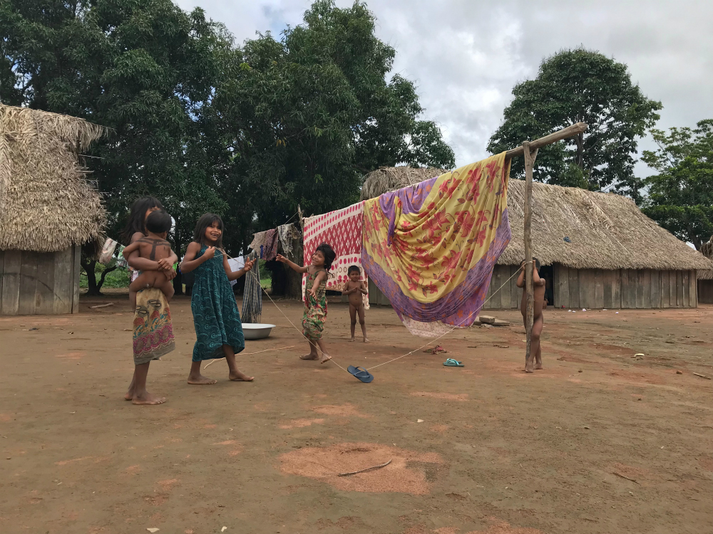 Five indigenous children in a village. Behind them are wooden houses with thatched roofs, and next to them is a clothesline with patterned sheets hanging on it. The children are playing with a long piece of string. A blue sandal hangs from the string. One of the children (in the left corner of the picture) wears a floral print dress. She is holding a baby in her arms.