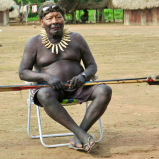 An indigenous man with short black hair, shirtless and wearing only green shorts, with his full body painted black from the nose down. He is sitting on a beach chair, with sunglasses resting on his head and holding a bamboo spear with a feather on its tip. The chair is on dirt ground, and behind him we see parts of a small wooden house with a thatch roof.